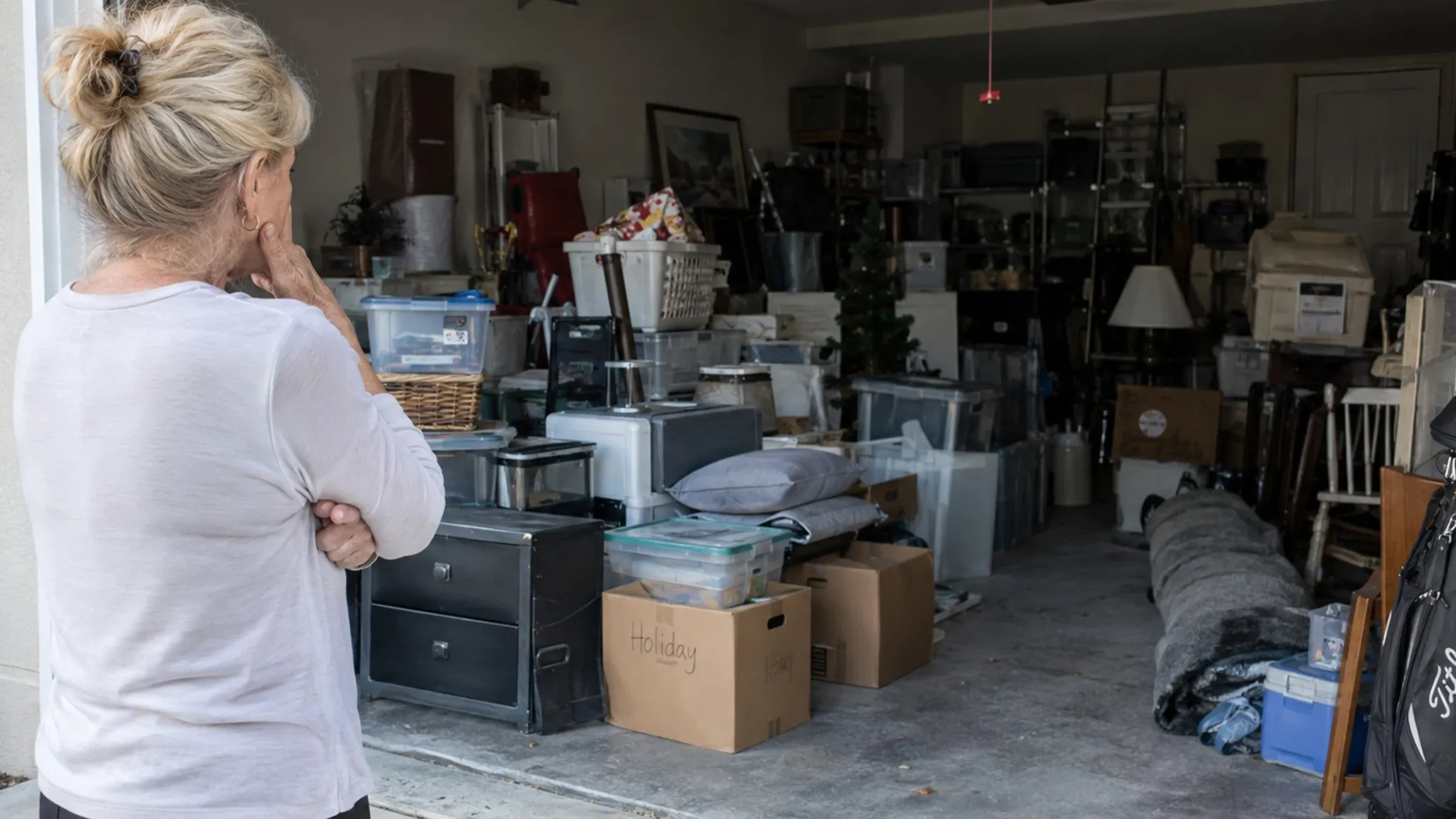 Homeowner standing in a driveway looking into a two-car garage packed to the ceiling with boxes, bins and stored furniture.